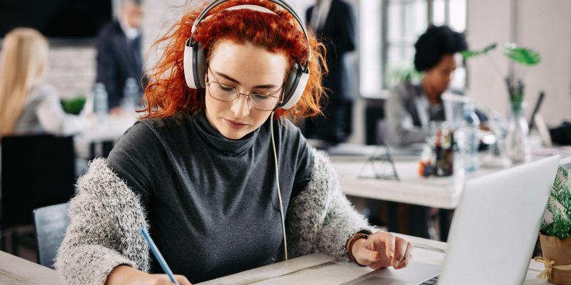 Redhead businesswoman using laptop and writing notes in her notepad while listening music on headphones at work.